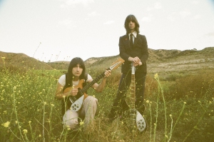 Two people with long hair in a field: one crouches holding a guitar, the other stands beside them with a guitar, wearing a suit. Sparse foliage and distant hills in the background.