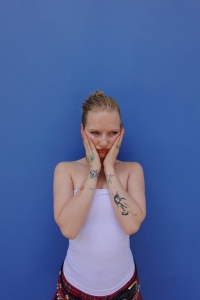 Person with tattoos holds their face with both hands, standing against a blue wall.