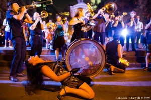 A brass band performs outdoors at night, with a person in the foreground energetically playing a large drum while leaning back.