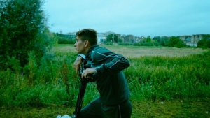 Person in a tracksuit leans on a scooter, looking out over a field with trees and distant buildings under a cloudy sky.