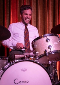 Person in a white shirt and patterned tie plays a drum set with a smile, against a backdrop of red and gold curtains.
