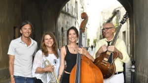 Four musicians stand in an archway: a man, a woman with a trumpet, a woman with an upright bass, and a man with a guitar. They are smiling at the camera.
