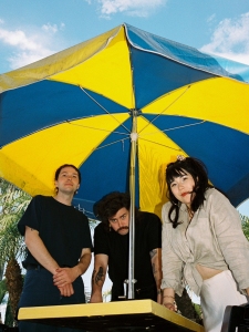 Three people stand under a large yellow and blue umbrella with a clear sky and palm trees in the background.