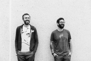 Two men stand against a textured wall, smiling and looking to the side. Both wear casual clothing, and one has tattoos. The photo is in black and white.