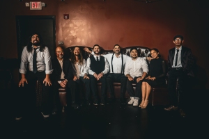 A group of eight people sitting on a couch against a dark background, wearing formal and semi-formal attire.