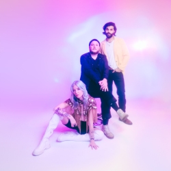 Three people pose against a soft, colorful background. One sits on the floor, and two stand behind. They are dressed in casual and semi-formal attire.