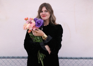 A person in a black dress holds a bouquet of colorful flowers, standing in front of a light-colored wall and a chain-link fence.