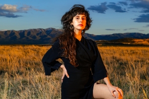 A person in a dark outfit stands confidently in a grassy field, with mountains and a cloudy sky in the background.