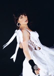 A person in a white dress with ribbon details holds a microphone against a dark background.