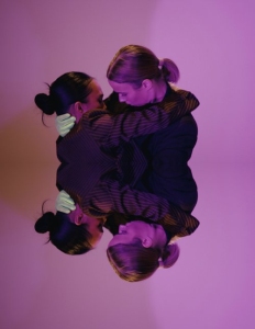Two people with their hair in buns embrace, surrounded by purple lighting, with their reflection beneath them.
