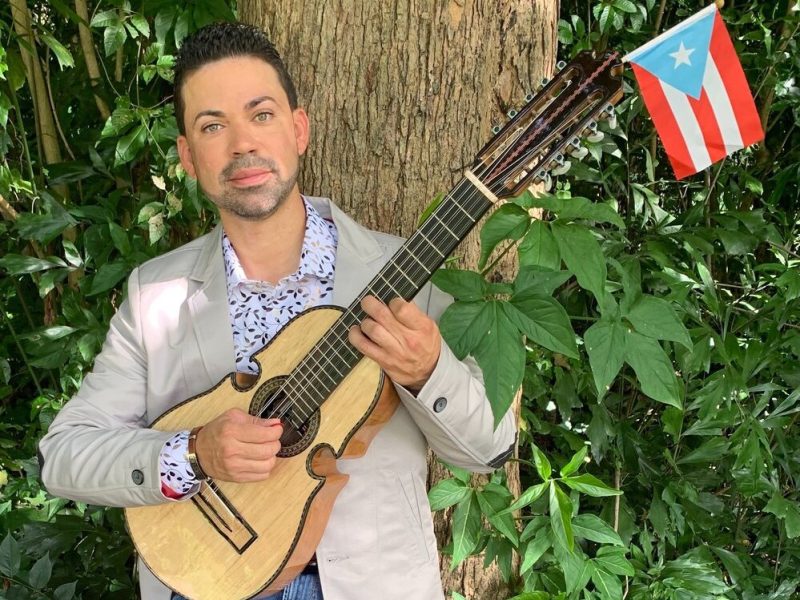 Person holding a guitar-like instrument adorned with a small Puerto Rican flag, standing in front of a tree surrounded by greenery.