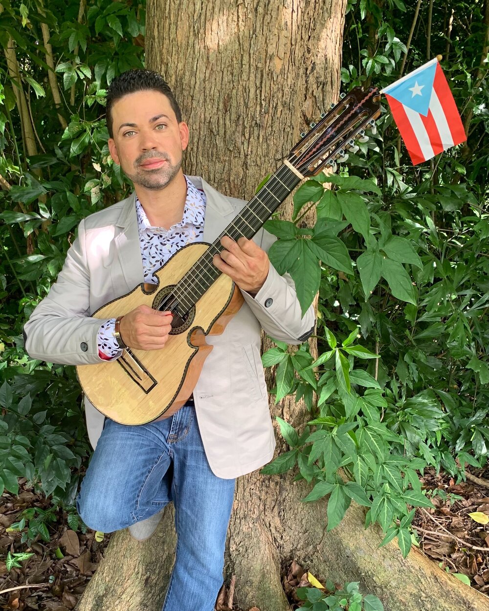 A man holding a guitar leans against a tree with a Puerto Rican flag nearby.