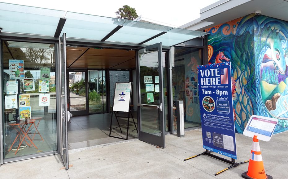 Entrance to a voting location with open glass doors and a sign displaying voting hours from 7am to 8pm. A traffic cone is nearby, and colorful murals are visible on the wall.