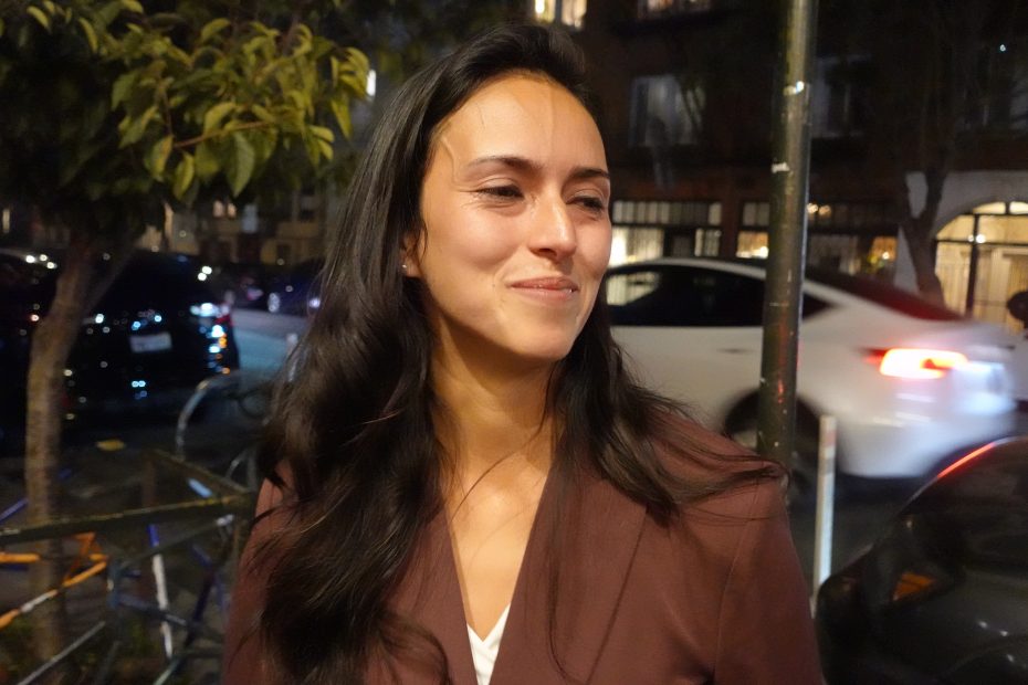 A woman with long dark hair smiles slightly, standing outdoors at night. Cars and trees are visible in the background.