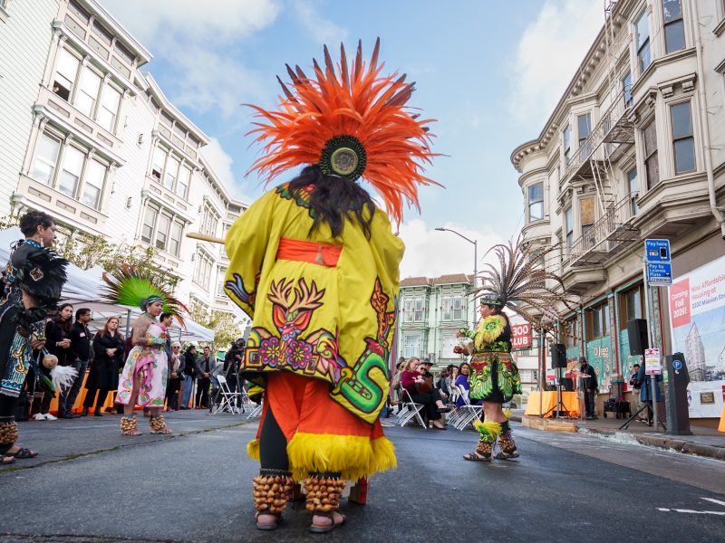 Performers in vibrant traditional attire and feathered headdresses dance on a street, surrounded by onlookers and historic buildings.