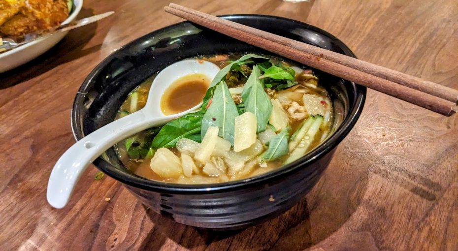 A black bowl of noodle soup with vegetables, garnished with basil and garlic, and accompanied by a spoon and chopsticks on a wooden table.