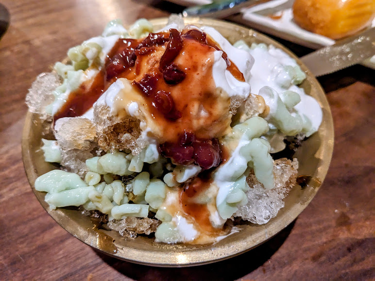 A bowl of dessert with crushed ice, green noodles, coconut milk, and red syrup, served in a metal bowl on a wooden table.