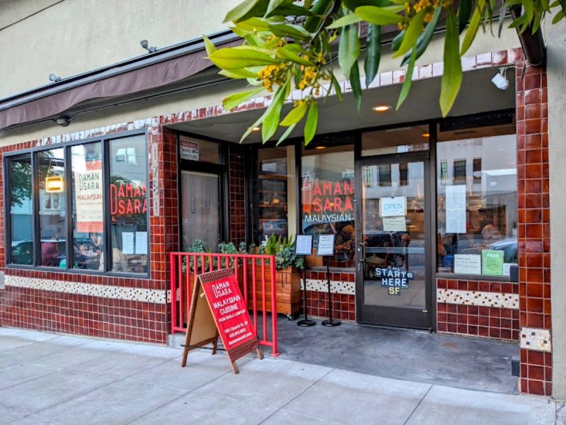 Street view of a Malaysian restaurant with tiled exterior, potted plants, and signage. A red menu board is placed outside near an entrance with a black "Start Here SF" sticker on the glass door.
