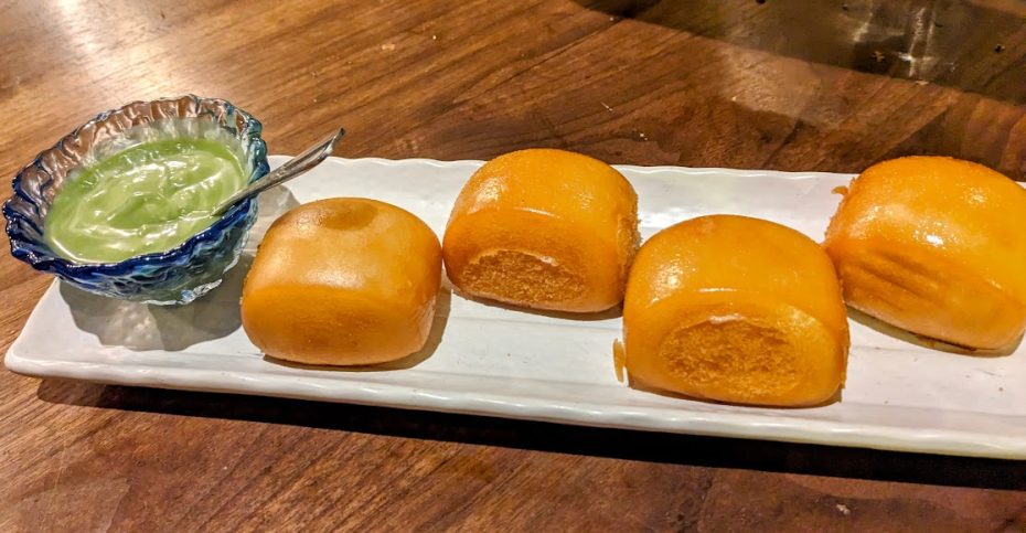 Four golden buns on a white rectangular plate with a small bowl of green dipping sauce on a wooden table.