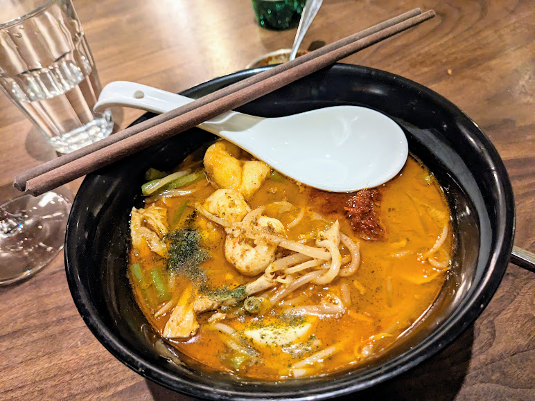 A bowl of spicy noodle soup with chopsticks and a spoon on top. A glass of water is on the wooden table beside it.