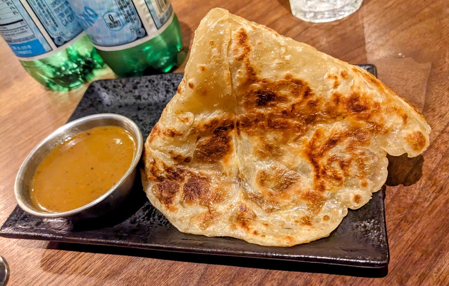 A plate with a piece of crispy flatbread alongside a small bowl of dipping sauce, placed on a wooden table.