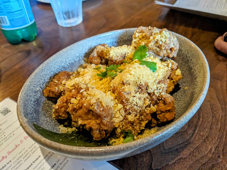 A bowl of fried chicken pieces garnished with grated cheese and cilantro, placed on a wooden table next to a menu and a bottle.