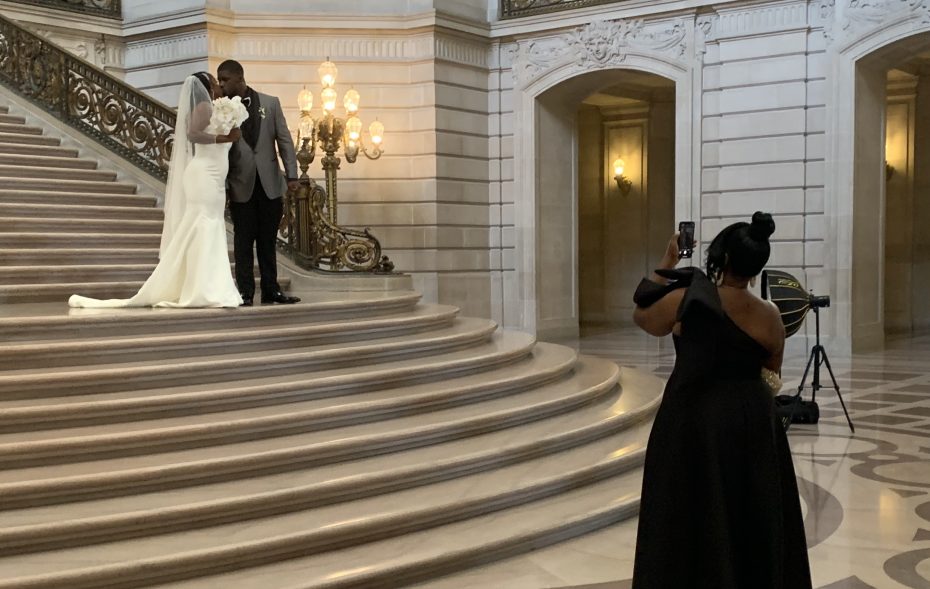 A couple poses for a photo on a grand staircase, while a person in a black dress takes their picture in an elegant, ornate interior.