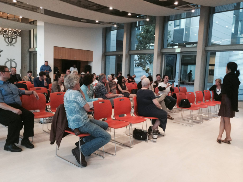 A group of people sit on orange chairs in a modern indoor setting with large windows, listening to a person speaking at the front.