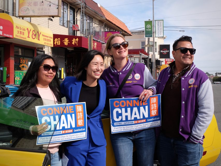 Four people stand together on a city street, smiling and holding campaign signs for Connie Chan.
