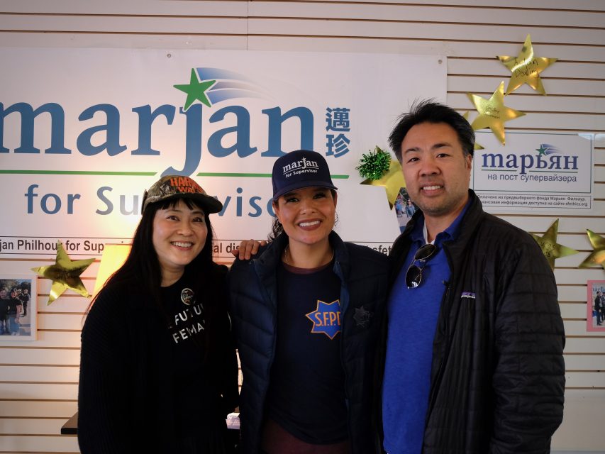 Three people standing in front of a campaign sign with stars and text. The two women wear hats with text, and the man wears a dark jacket.