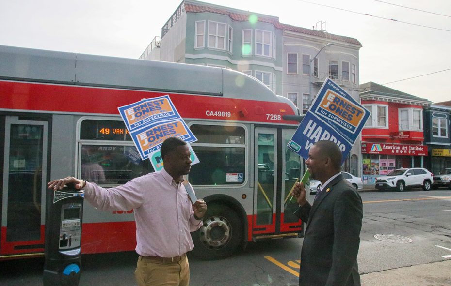Two people on a sidewalk hold campaign signs for "Ernest Jones" and converse near a passing city bus in an urban street setting.