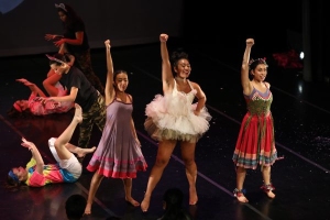 A group of performers on stage, with three standing in front, raising one arm, while others perform on the floor in the background.