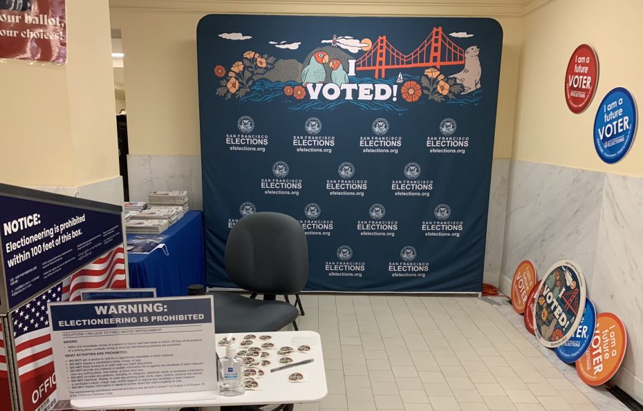 Voting station with "I Voted" backdrop, chair, and table displaying pins and a sign warning against electioneering. Posters and informational materials are visible in the background.