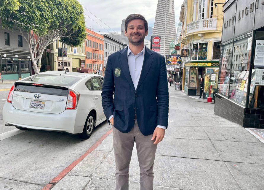 A person in a blazer and pants stands on a city sidewalk near parked cars and buildings.