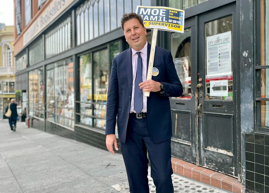 Person in a suit holding a campaign sign on a city sidewalk in front of a building with glass windows.
