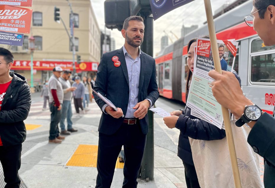 A man in a suit holds a clipboard and talks to people on a city street. Banners and a bus are visible in the background.
