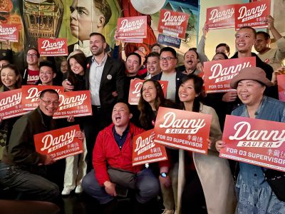 A group of people smiling and holding "Danny Sauter for D3 Supervisor" signs, gathered in a lively indoor setting.