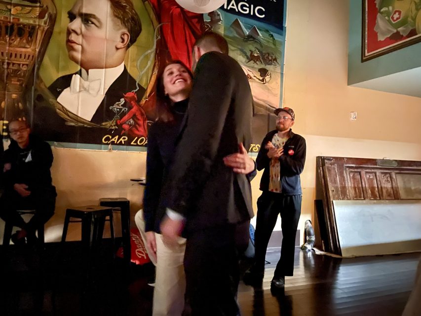 A couple embraces in front of a vintage poster, while a man stands nearby. Indoor setting with wooden floor and stools.