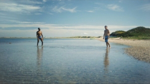Two people stand in shallow water at a beach, with a clear sky and greenery on the horizon.