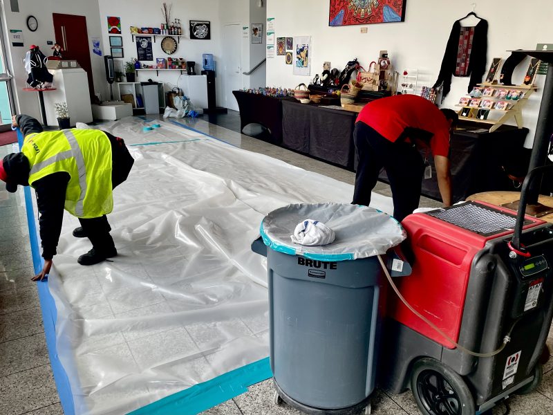 Workers lay plastic sheeting and tape on a tiled floor inside a room with various items displayed and a large trash bin in the foreground.