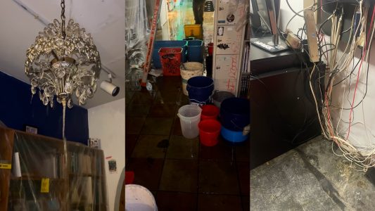 A cluttered room with a leaking chandelier, buckets collecting water, and exposed wires on the floor, indicating a damaged ceiling and water leak situation.