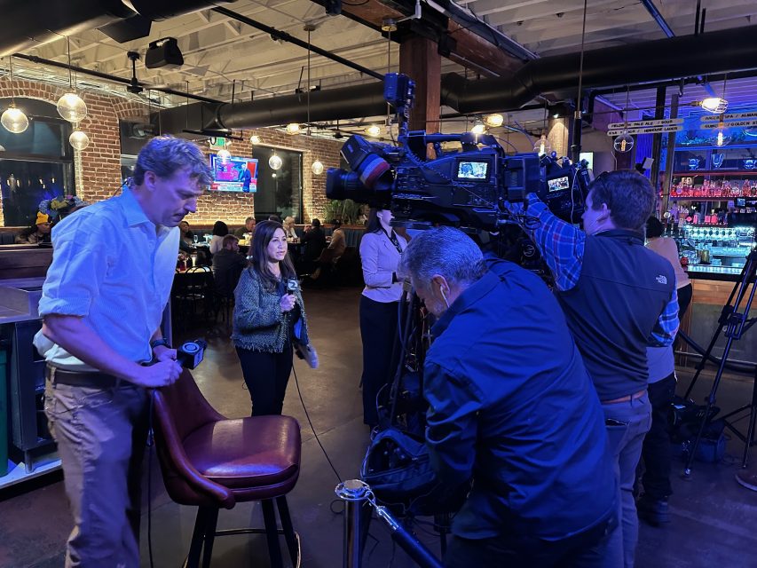 A news crew sets up cameras in a bar with patrons in the background. A man places a chair, and a woman stands nearby preparing for a broadcast.