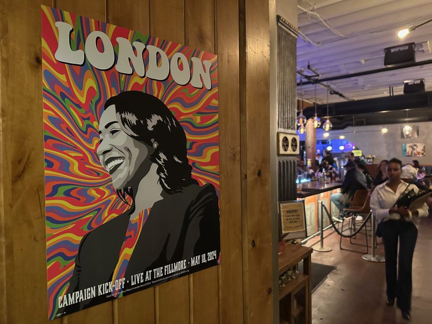 Colorful poster on a wooden wall with a person smiling, labeled "London," advertising a campaign kickoff event at the Fillmore on May 8, 2024. Background shows a bar with people.