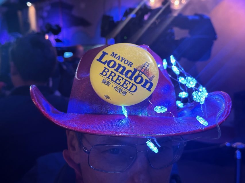 A person wearing a hat with a "Mayor London Breed" sticker and decorative lights.