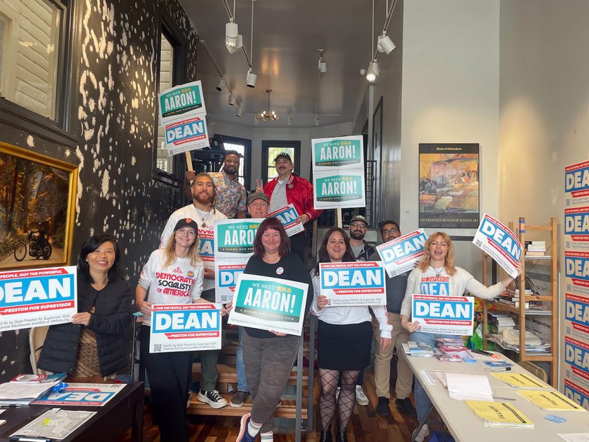 A group of people indoors holding campaign signs that say "Dean" and "Aaron!" in a brightly lit room.