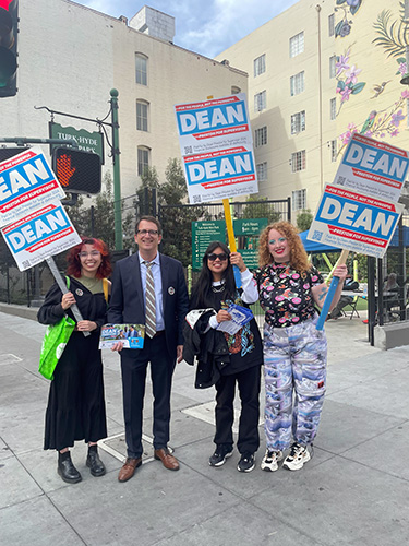 Four people on a city street hold campaign signs that say "Dean" while smiling at the camera.