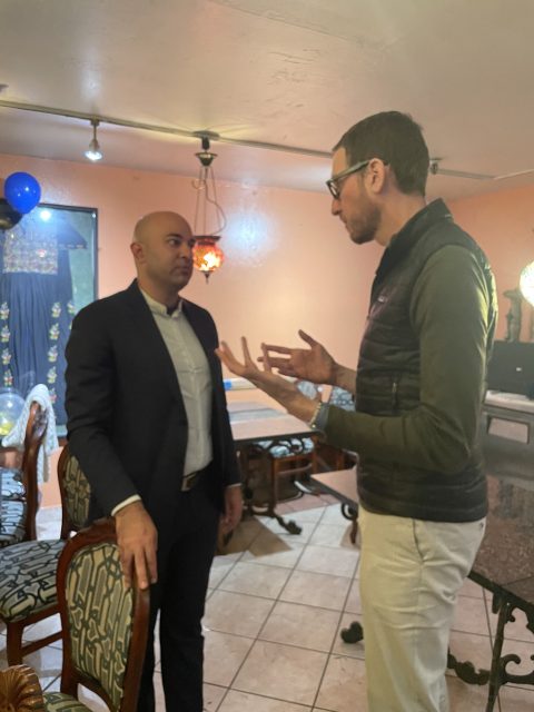 Two men are having a conversation in a dimly lit room with orange walls. There's a chair and table nearby, and balloons are visible in the background.
