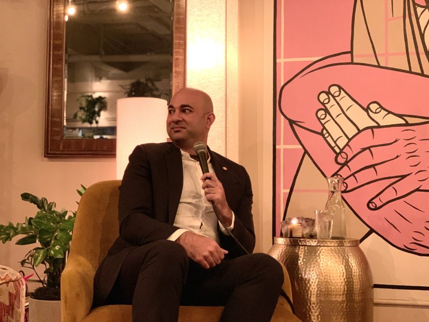A man in a suit sits on a yellow chair holding a microphone, with a plant and a small table beside him, in a room with a large artwork on the wall.