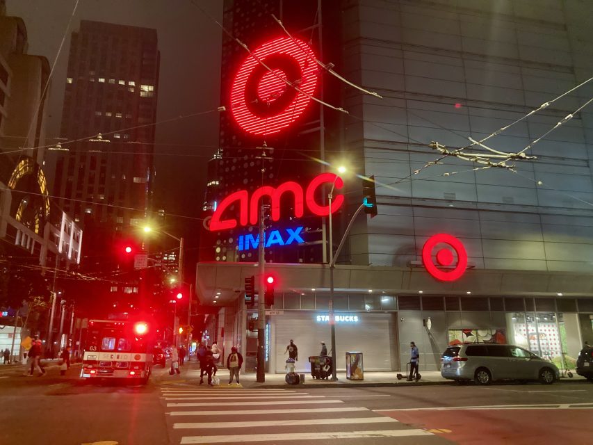 Nighttime city scene with AMC IMAX, Target, and Starbucks signs on a building. People walk across a street intersection, and a fire truck is parked nearby.