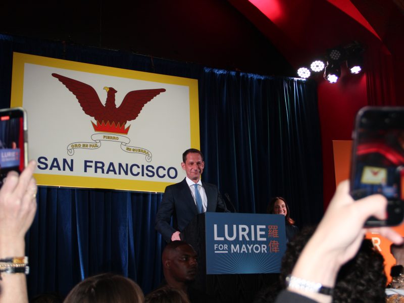 Person speaking at a podium labeled "Lurie for Mayor" with people holding phones. A large San Francisco emblem is displayed in the background.